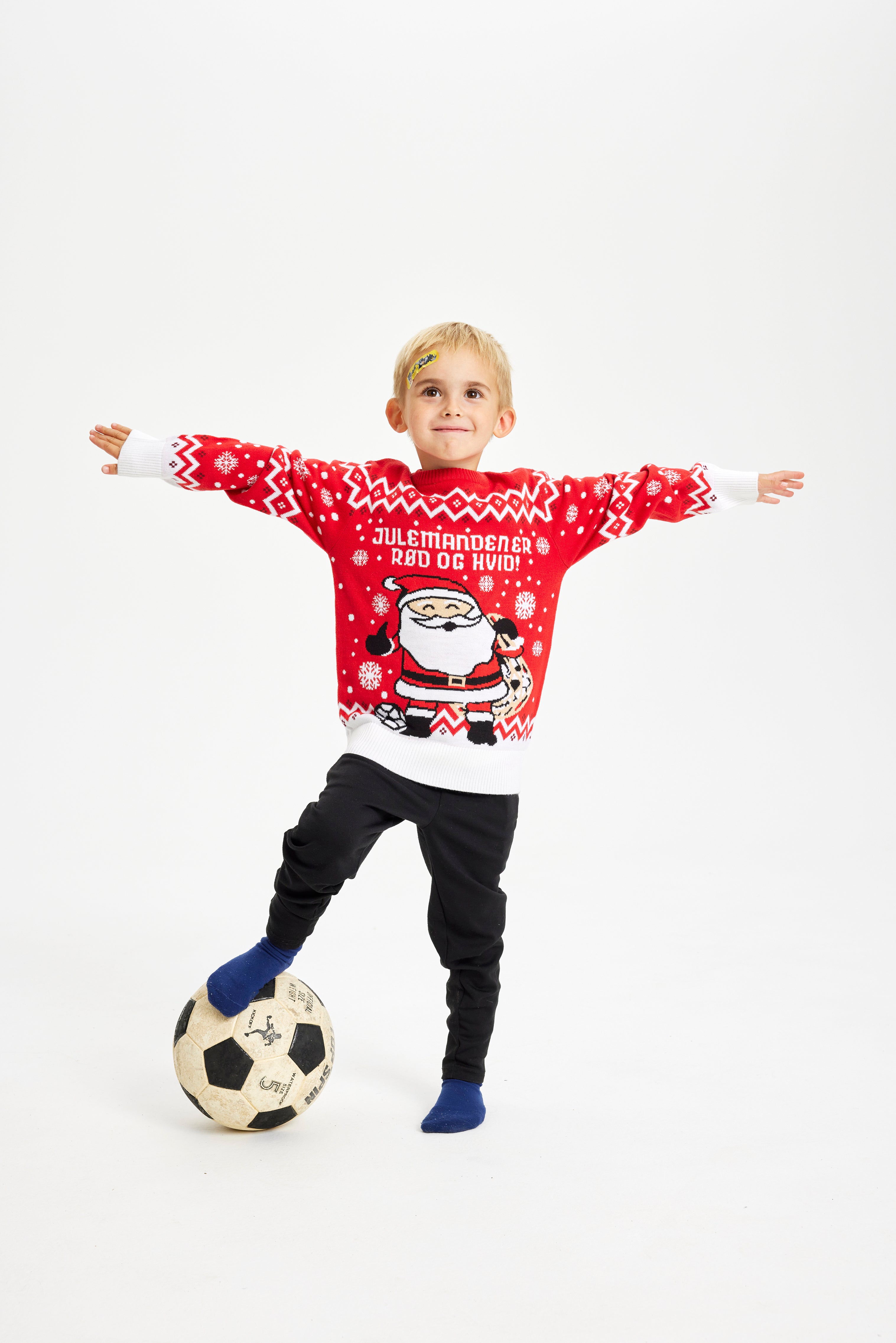 Et barn iført en rød julesweater står med en bold under den ene fod.