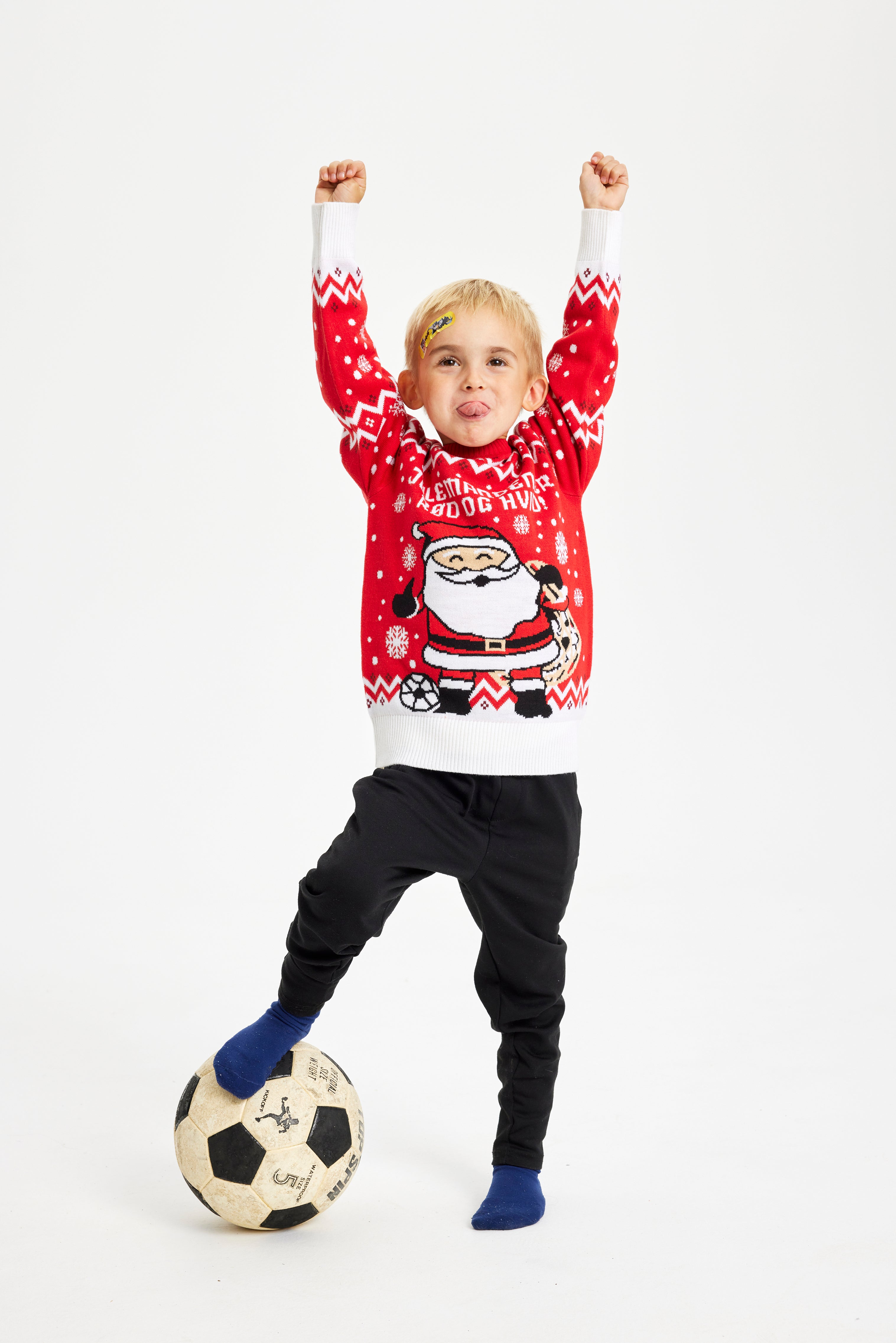 Et barn med hænderne i vejret står iført en rød julesweater og med en bold under den ene fod.