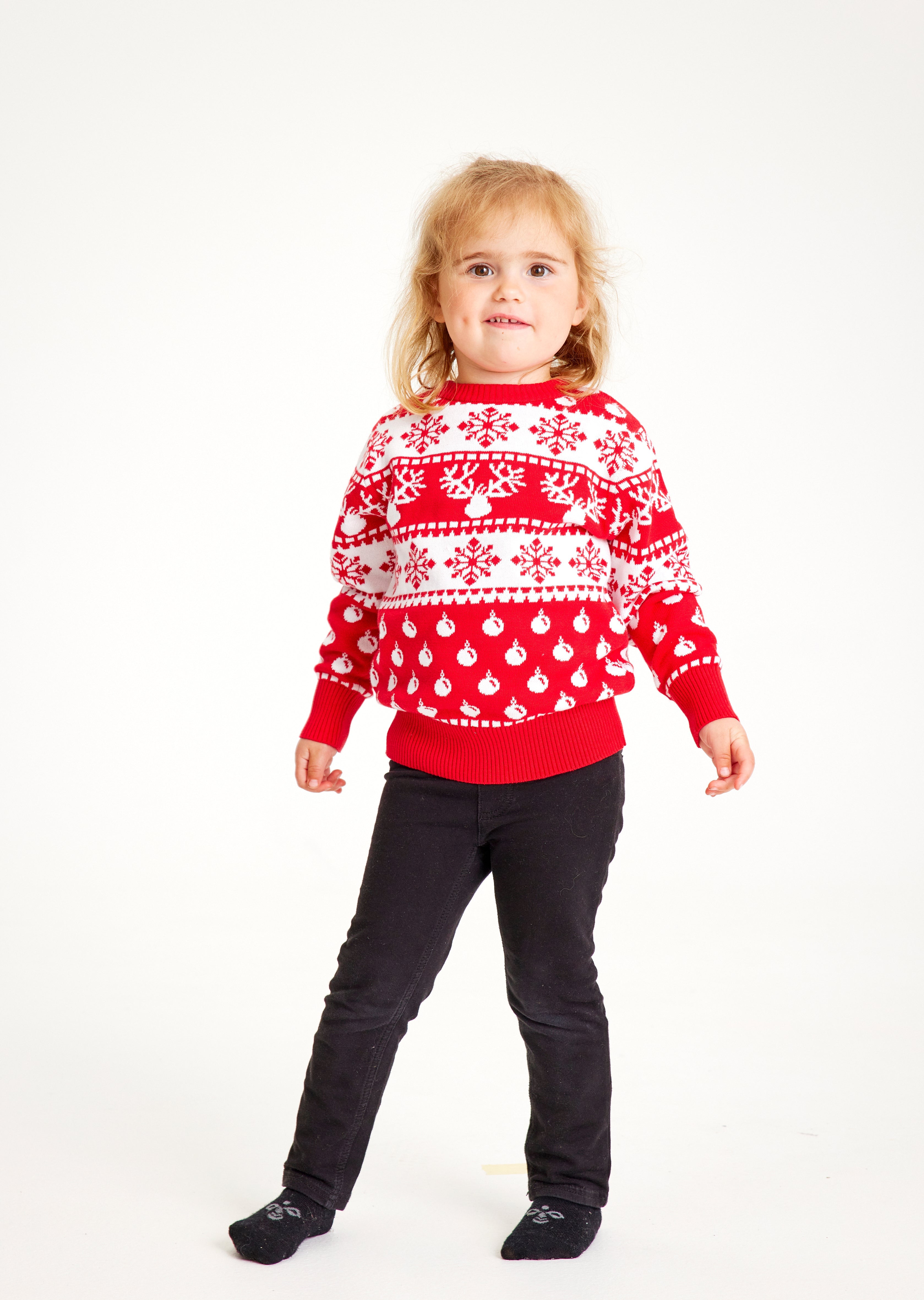 Et barn poserer i en rød julesweater med julekugler, rensdyr og snefnug.