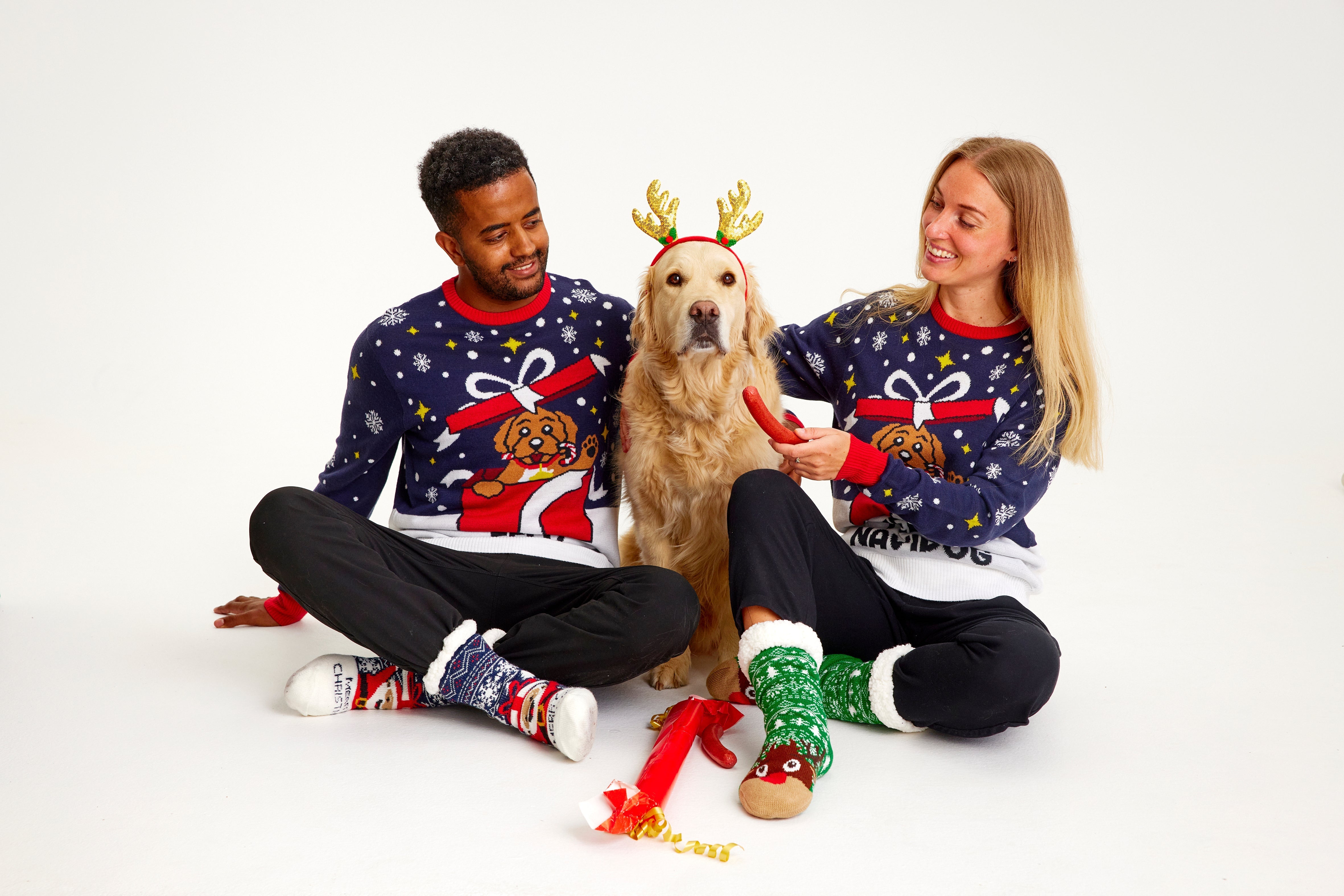 En mand og en dame iført hver deres julesweater sidder sammen med en hund.